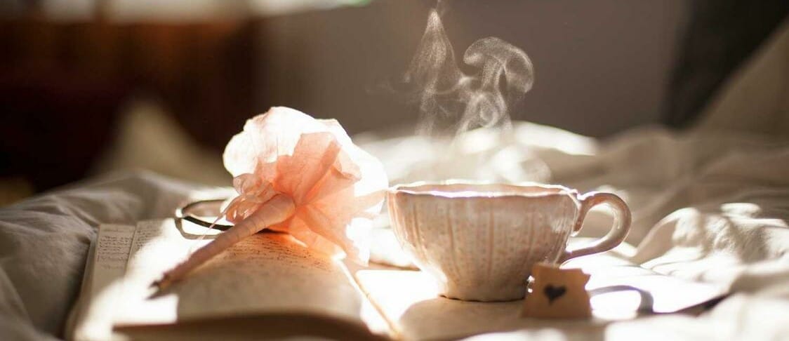 A pen, notepad and mug filled with a steaming hot drink resting on a soft surface