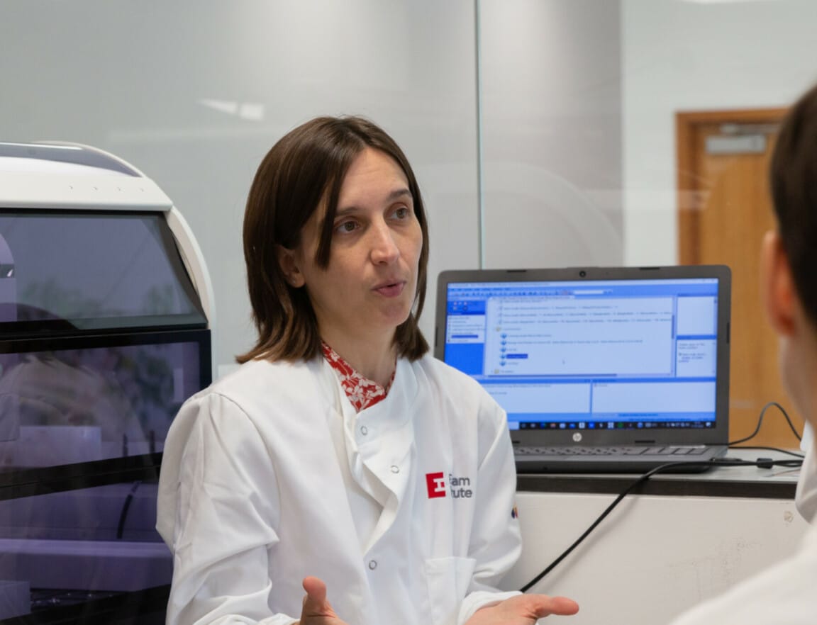 A person in a white lab coat speaking to a small group of people in a laboratory