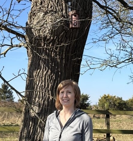 Dr Gabrielle Davidson standing outside next to a large tree with a brown and pink bird box