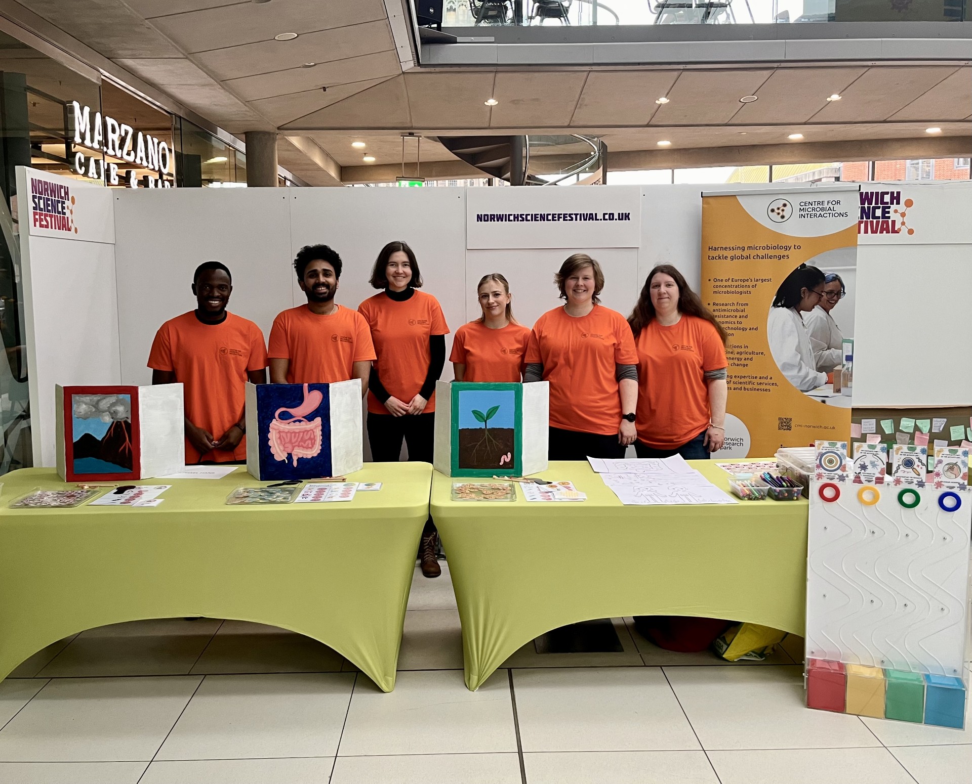 A group of volunteers wearing bright orange t-shirts, standing in front of a tabletop stand ready to deliver outreach activities at Norwich Science Festival.