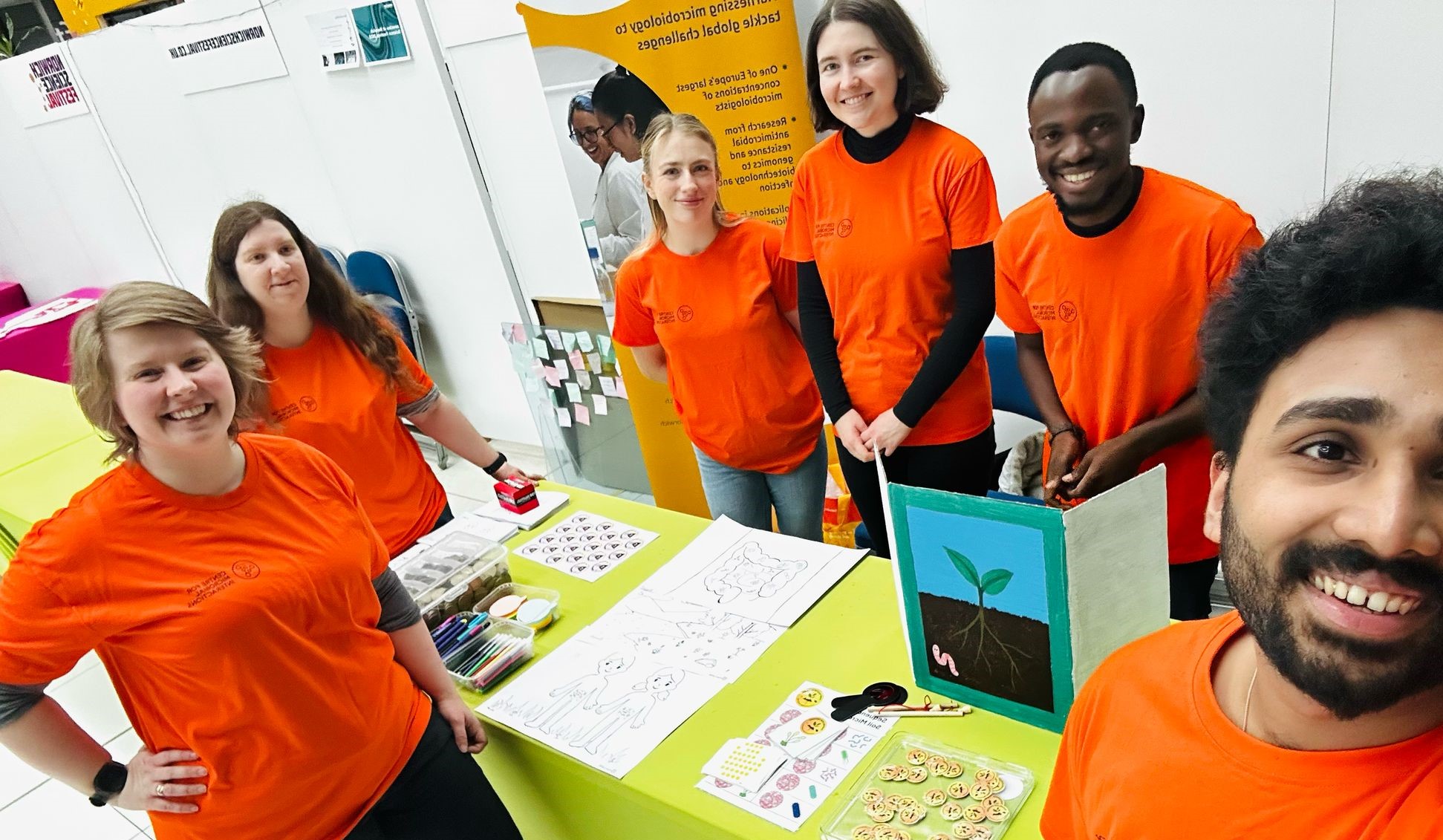 A group of volunteers wearing bright orange t-shirts, standing around an activity stand for the public at Norwich Science Festival