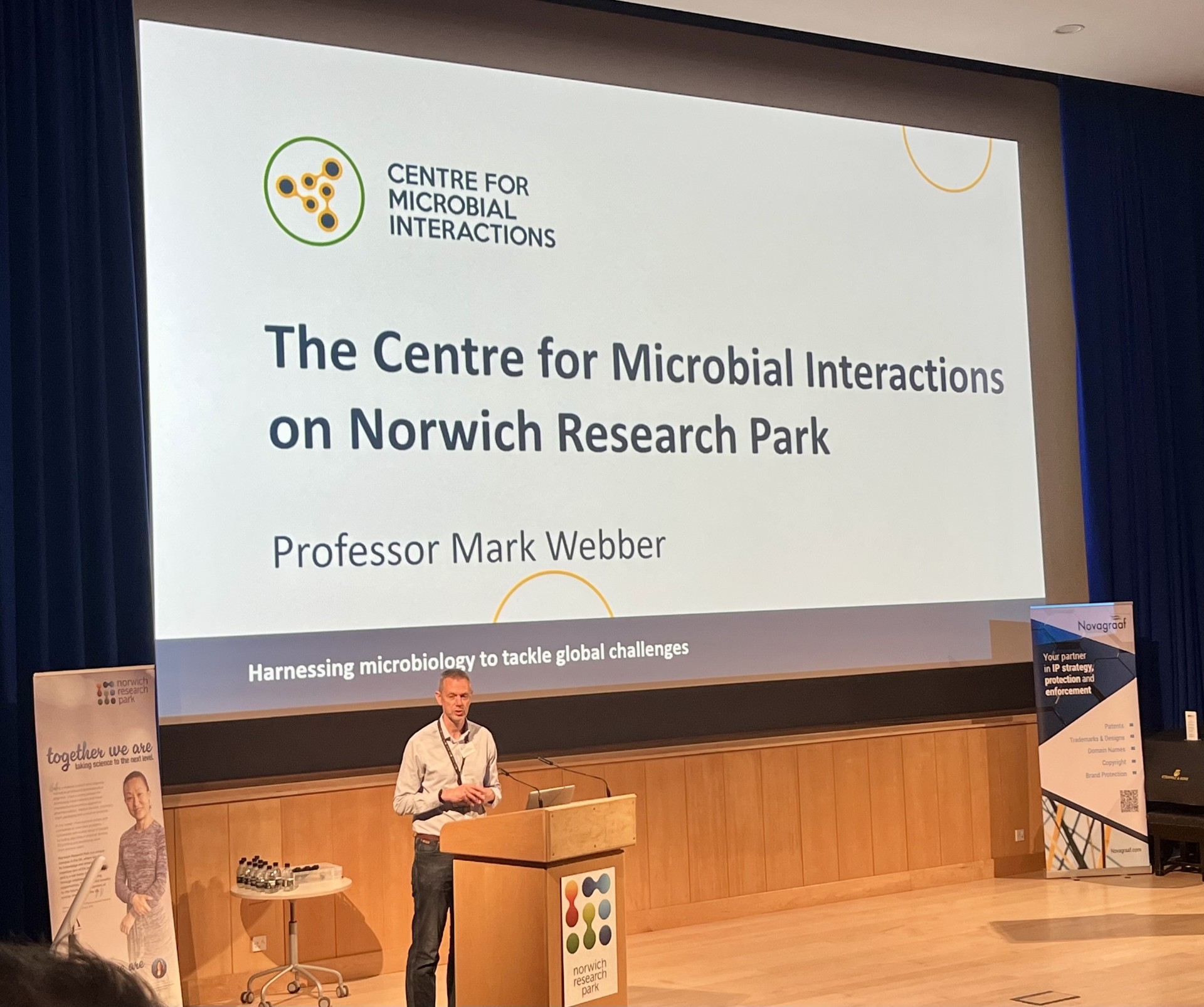 Professor Mark Webber presenting about the Centre for Microbial Interactions from a raised conference stage in front of a large screen