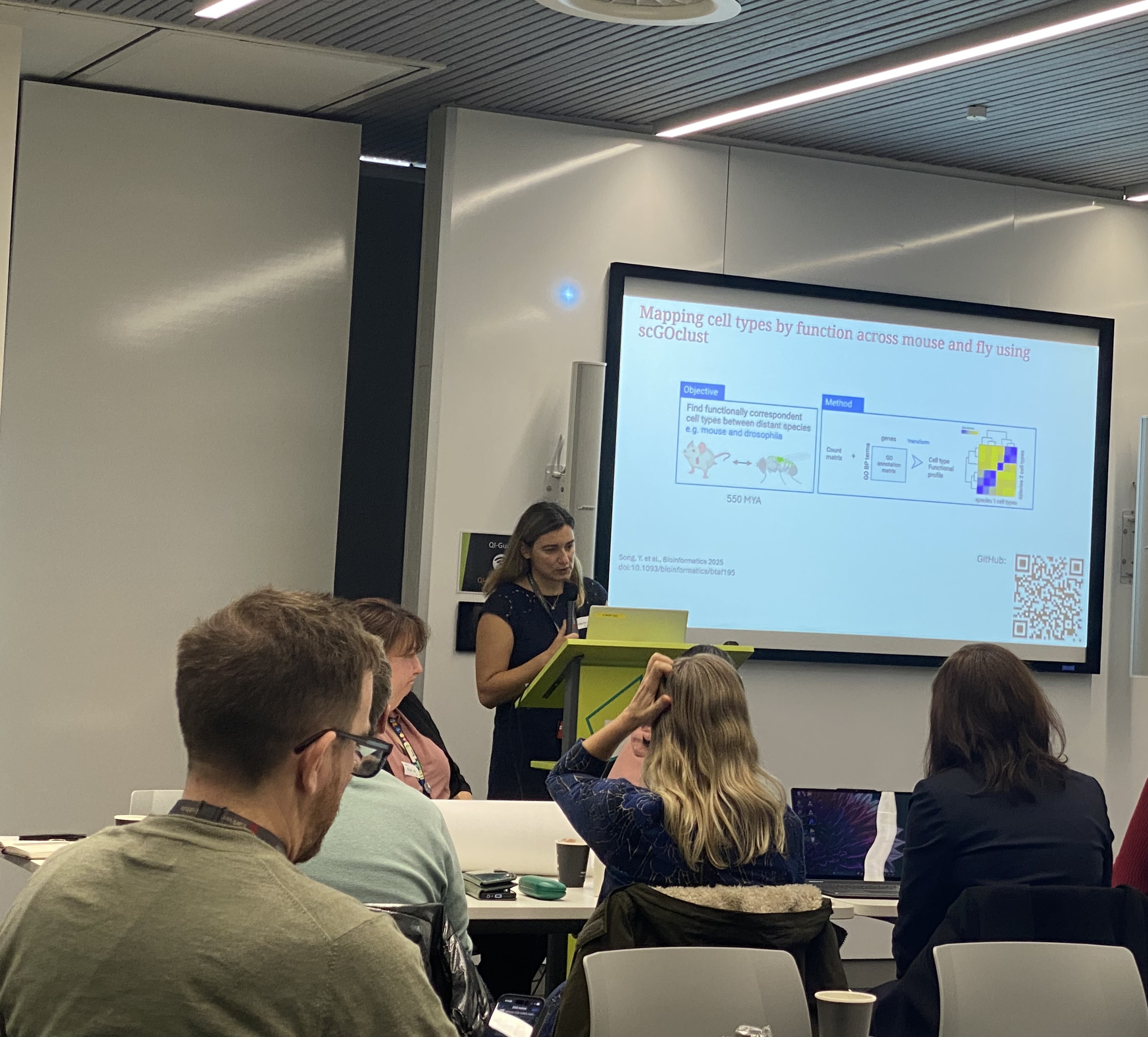 Irene Papatheodorou standing in front of a large presentation screen and sharing research with symposium attendees