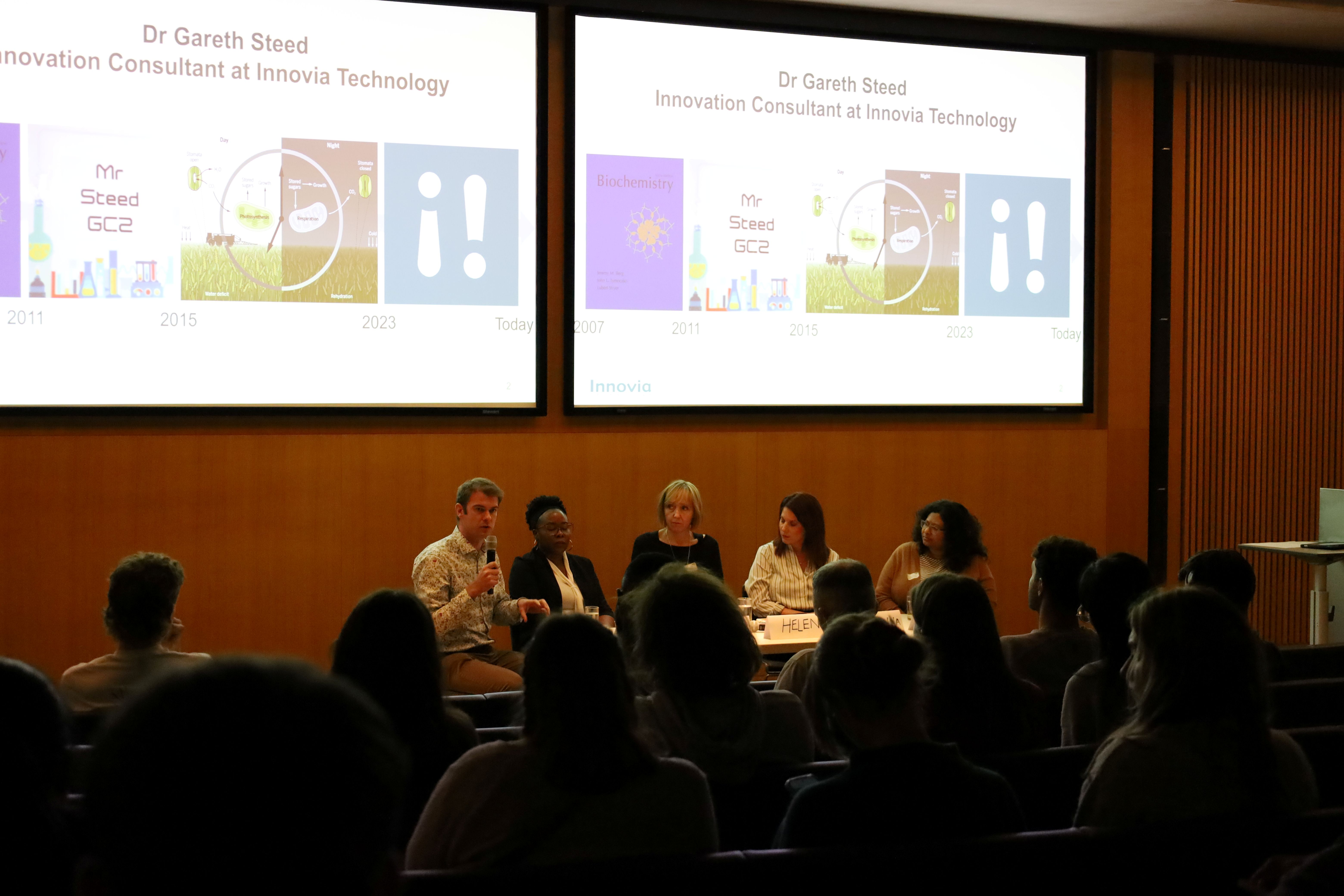 Five panel members speaking to a large audience from the front of a lecture theatre.