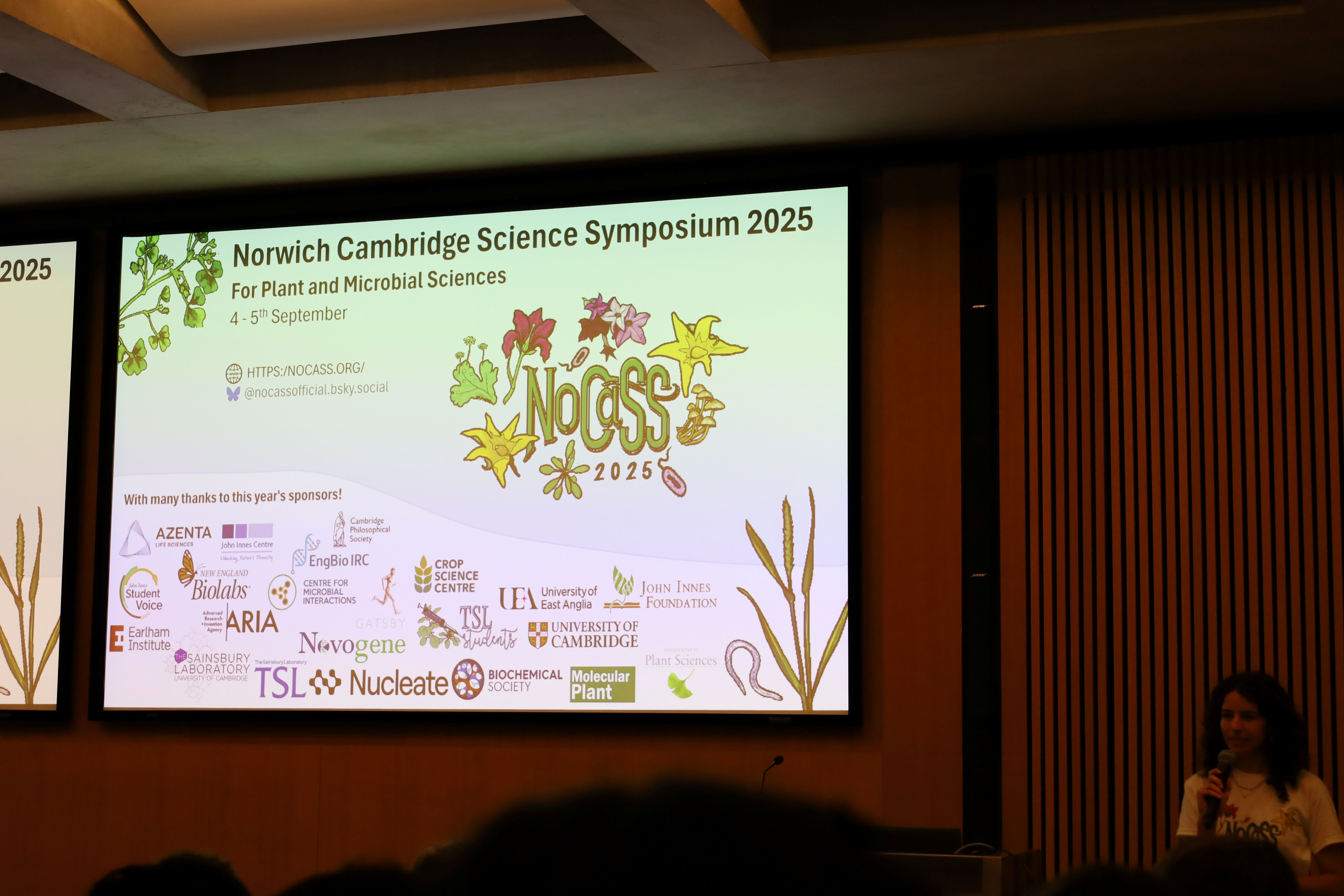 Someone presenting in front of a large screen showing the NoCaSS logo and sponsor logos