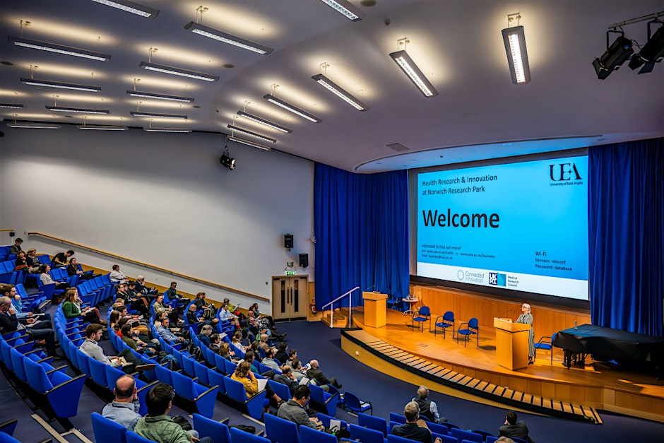 A large lecture theatre with attendees watching a presentation on a big digital screen