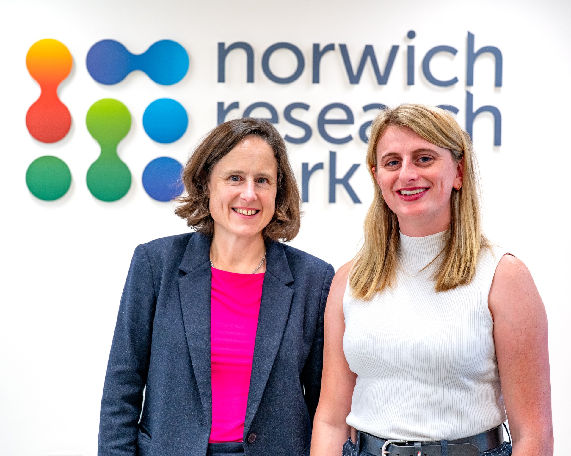 Roz Bird and Dr Alicia Showering standing in front of the Norwich Research Park logo