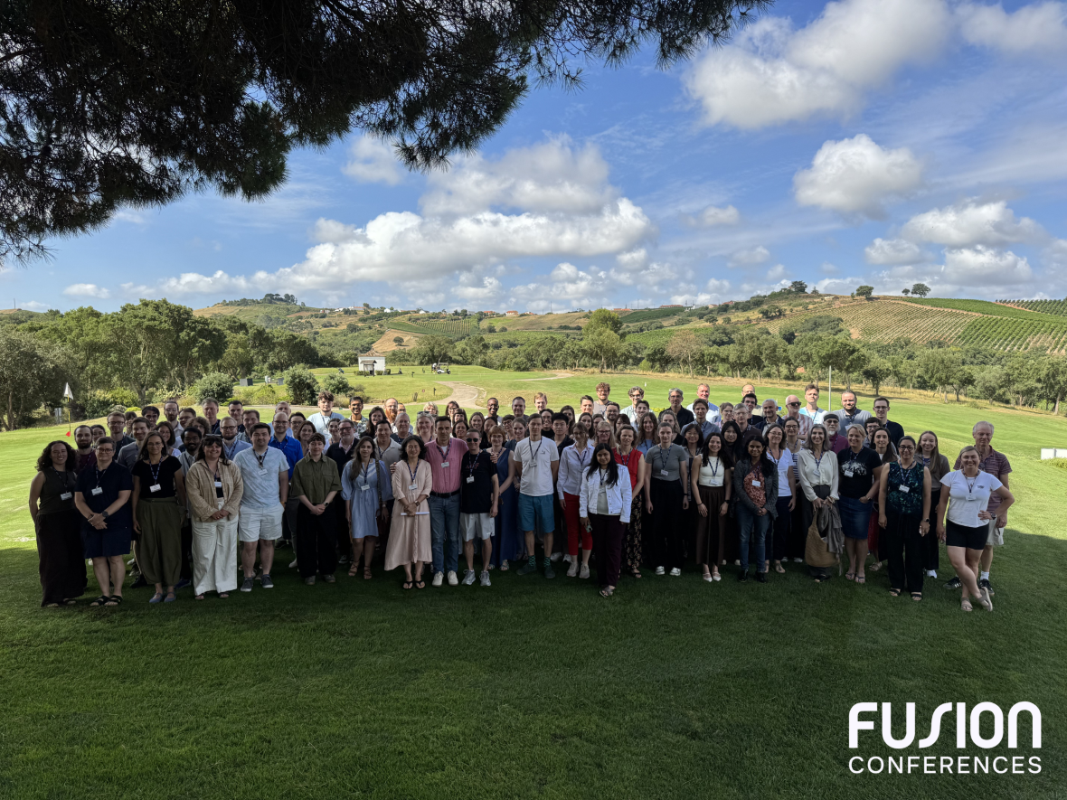 A group of around 100 conference attendees standing together outside. The sky is blue and slightly cloudy with green hills in the background.