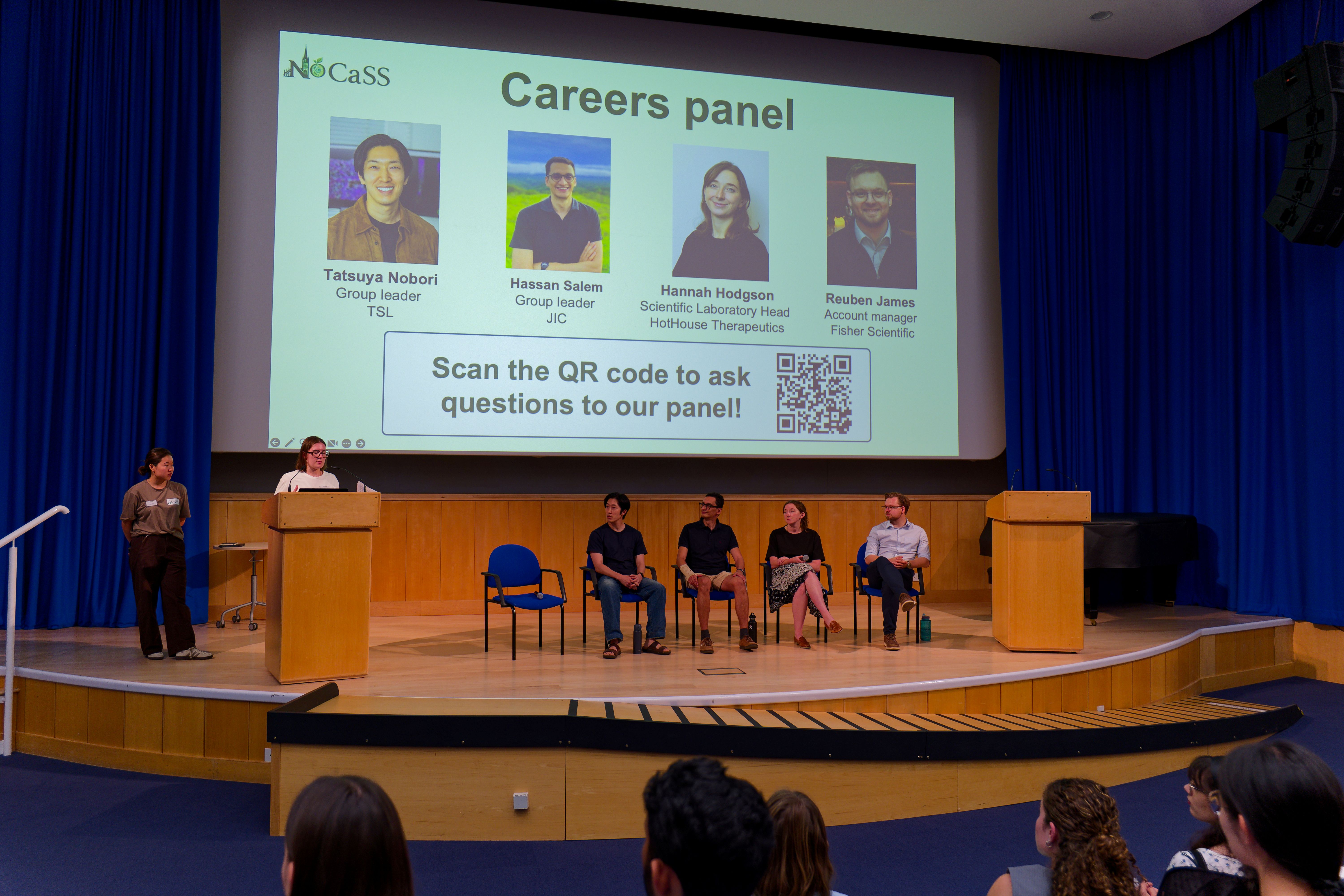 Careers panel at the conference with four speakers sitting on chairs in front of a large presentation screen and another speaker to the left behind a microphone podium
