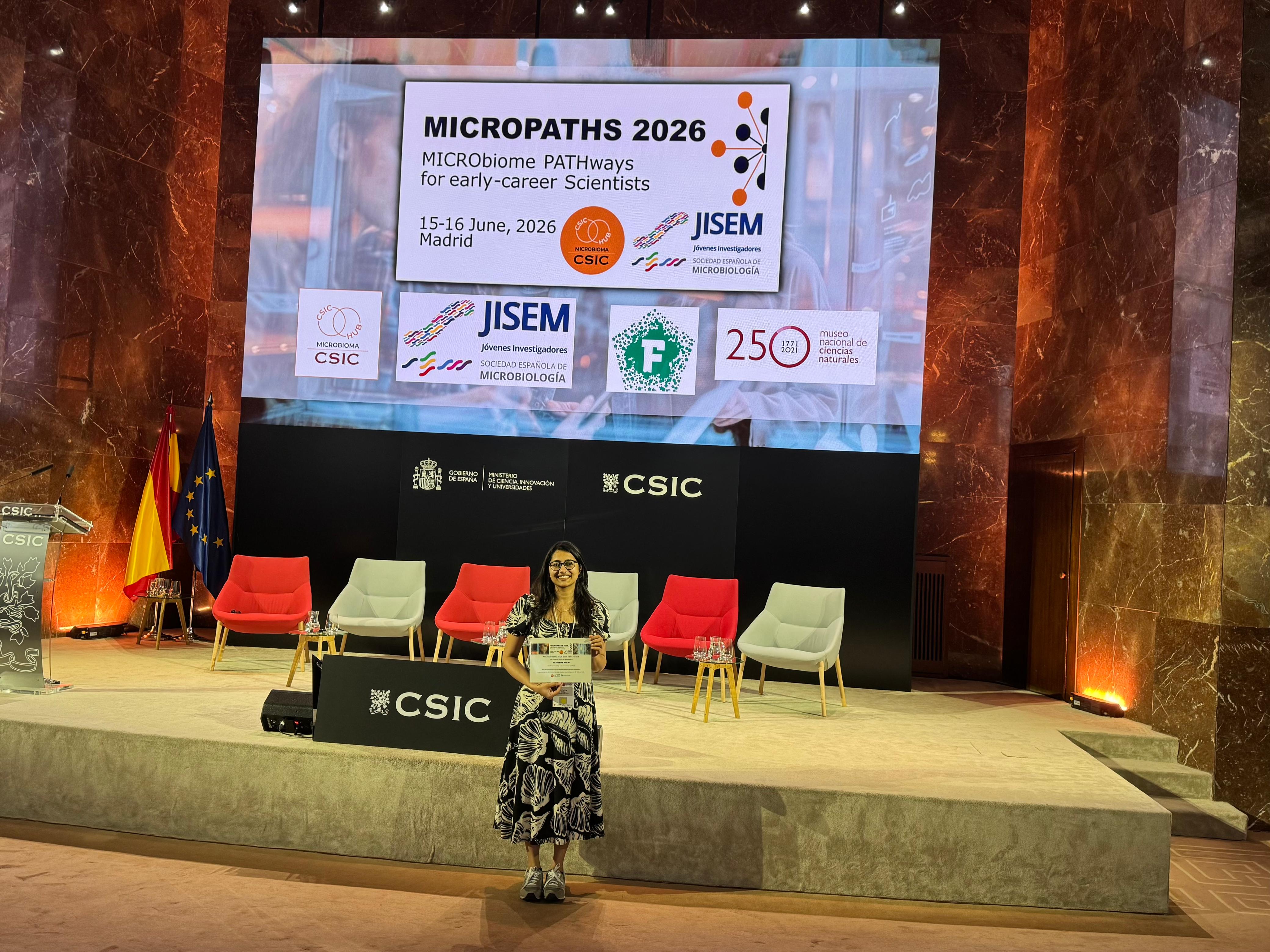 Catherine Philip holding a poster prize certificate standing in front of a stage and large digital screen